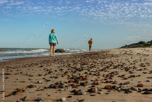 Barefoot careless man and woman walking on pebble sandy beach on sunny summer day.People searching for amber stone in Baltic Sea.Travelers enjoying vacation.Holiday travel meditation relax concept