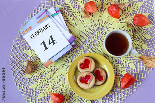 Calendar for February 14: the name of the month February in English, the numbers 14 on the loose-leaf calendar, a cup of tea, heart-shaped cookies, physalis branches on a yellow openwork napkin