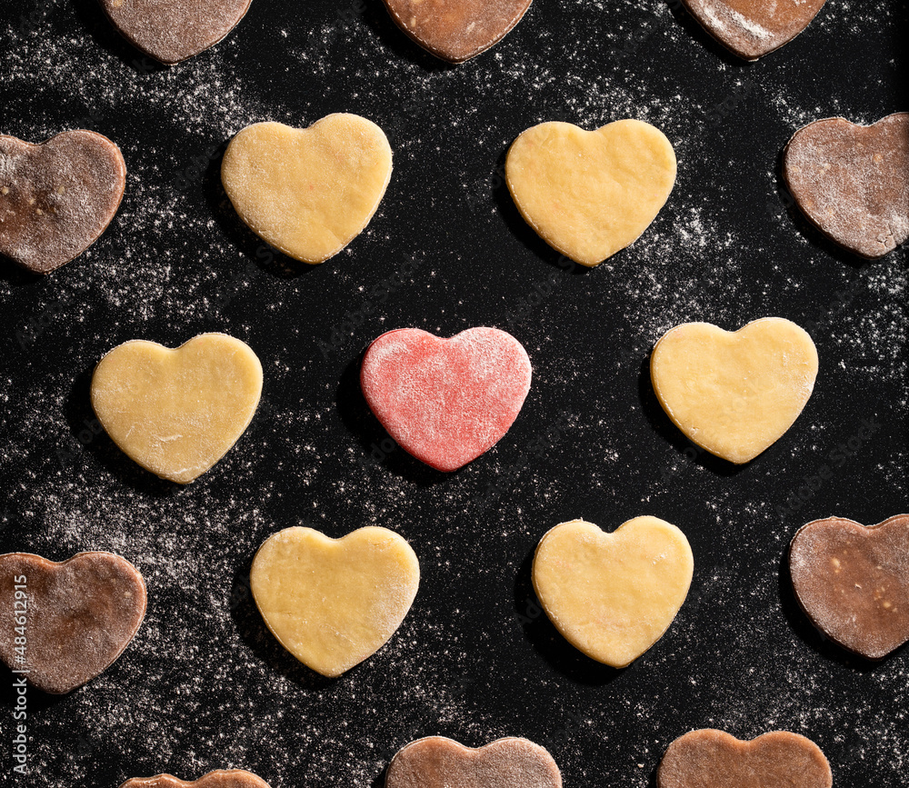 Macro view of pre-baked vanilla, pink and chocolate cookies in shape of hearts dusted with flour on black baking sheet