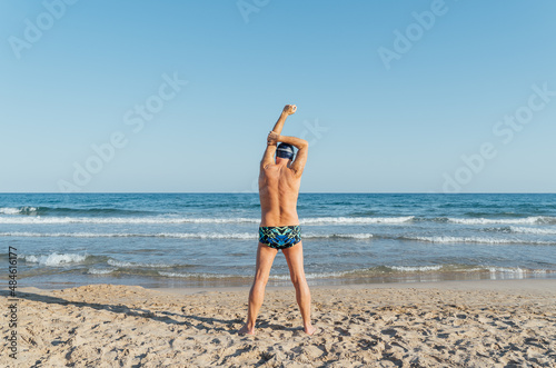 Senior swimmer stretching with his back to the camera before entering the sea. Training on the beach