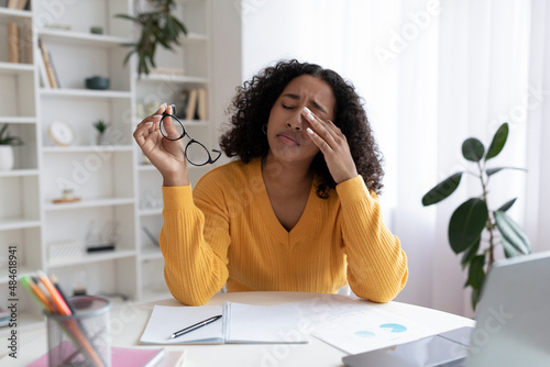 Fotografia Young black woman feeling tired, rubbing irritated eyes, sitting at desk with la