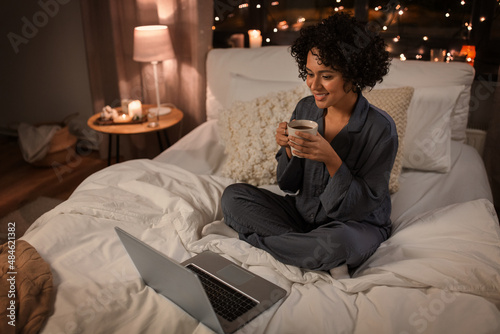 technology, bedtime and rest concept - happy smiling woman in pajamas with laptop computer and mug sitting in bed at night and drinking coffee
