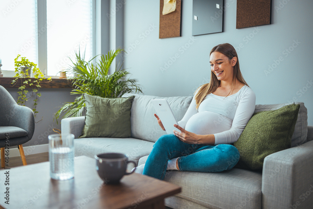 Attractive young pregnant woman smiles while using her digital tablet to video chat while relaxing in her living room. Pregnancy, technology, people and expectation concept Attractive young pregnant woman smiles while using her digital tablet to video chat while relaxing in her living room. Pregnancy, technology, people and expectation concept