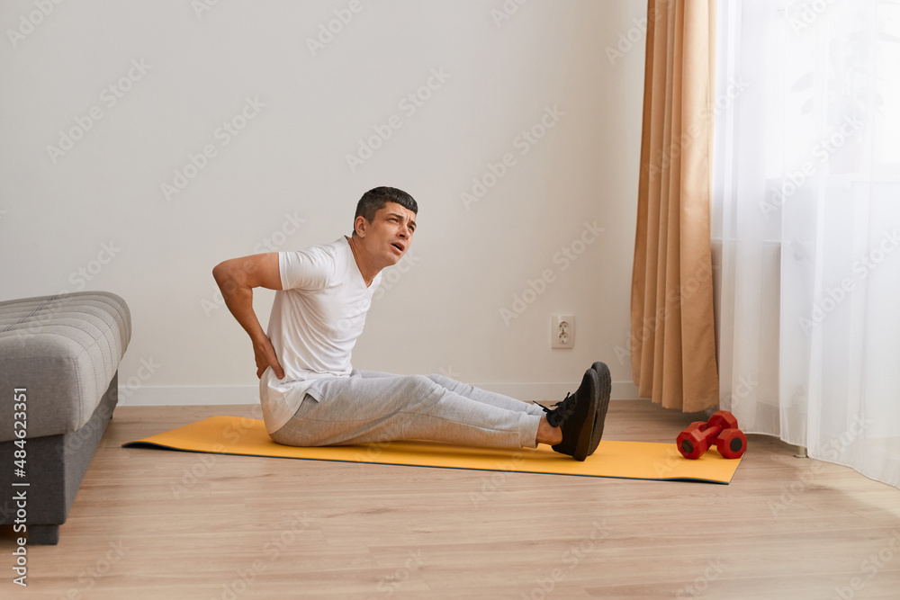 Portrait of young adult handsome sporstman sitting on mat doing stretching yoga exercise at gym, suffering of backache, touching back with hand, muscular pain.