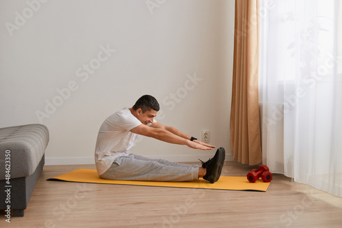 Indoor shot of sporty athletic man wearing sportswear sitting on yoga mat on floor at home and doing stretching, having workout, training flexibility.