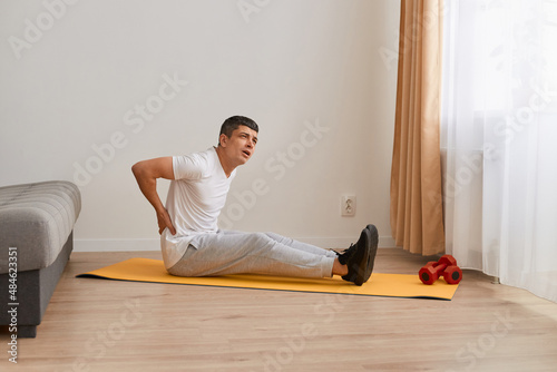 Portrait of young adult handsome sporstman sitting on mat doing stretching yoga exercise at gym, suffering of backache, touching back with hand, muscular pain.