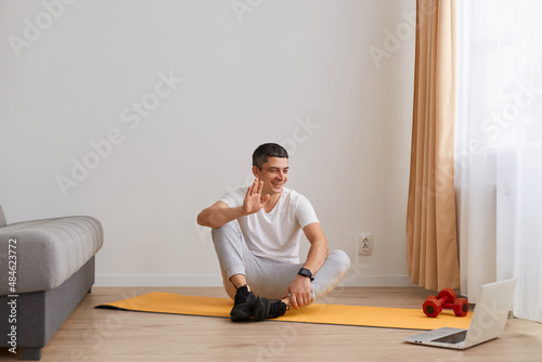 Smiling young adult brunette man sitting on mat on floor and waves hand to laptop webcam in living room interior, healthy lifestyle, online class, workout at home during.