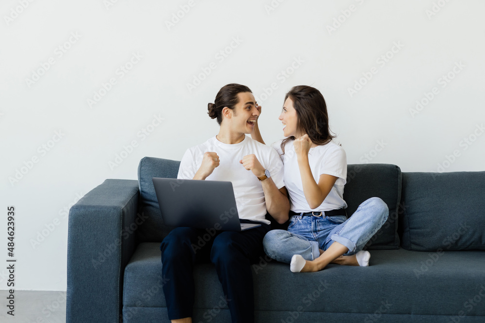 Overjoyed excited couple getting good news, celebrating success, goal achieving, winning prize. Happy man and woman sitting on couch with laptop, making winner gesture, shouting for joy