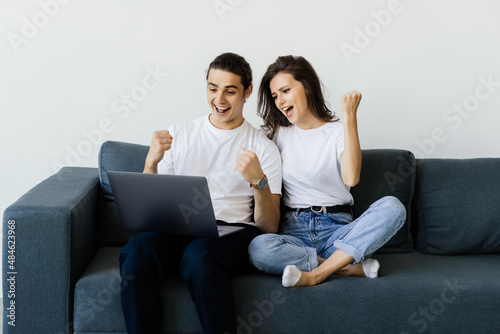 Overjoyed excited couple getting good news, celebrating success, goal achieving, winning prize. Happy man and woman sitting on couch with laptop, making winner gesture, shouting for joy