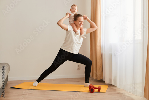 Portrait of sporty female doing yoga practice on yoga mat at home with baby daughter on her shoulder, healthy lifestyle, fitness, sport exercises near window i living room.