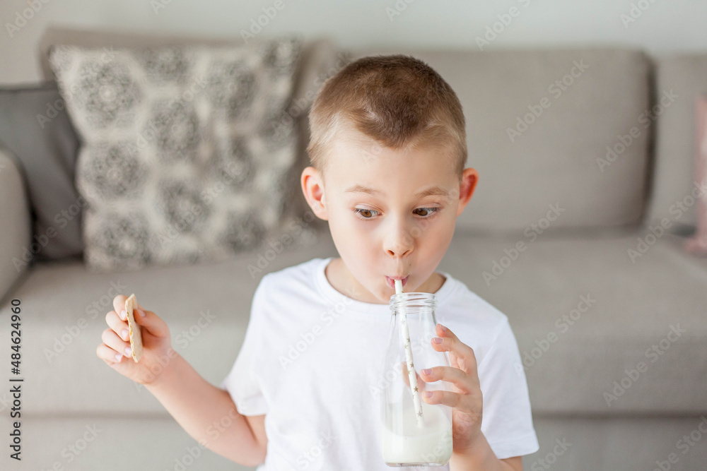 Adorable happy fair-haired boy in light clothes drinks milk from a glass bottle and eats a loaf of bread at home. Eco products. Healthy food and drinks. Adorable happy fair-haired boy in light clothes drinks milk from a glass bottle and eats a loaf of bread at home. Eco products. Healthy food and drinks.