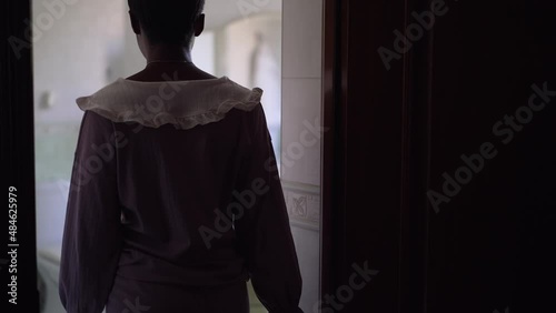 Young slim African American woman in pajamas entering bathroom sitting down on pouf smiling. Portrait of happy relaxed beautiful lady at home indoors in the morning. Slow motion