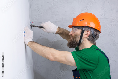 An electrician installs sockets in the apartment. A guy in an orange helmet and overalls makes electrics in the house.