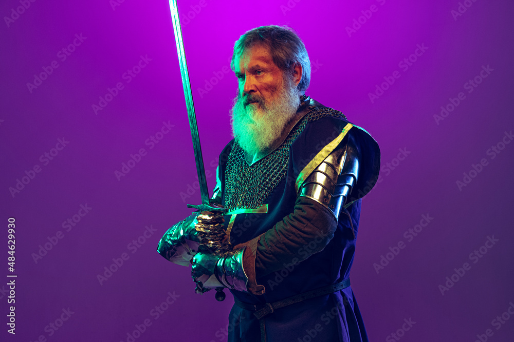 Portrait of senior greybearded man, brave and brutal medieval warrior or knight in armor with sword isolated over purple background in neon. Comparison of eras