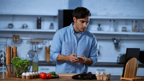 young man holding smartphone and looking at vegetables on table in kitchen.