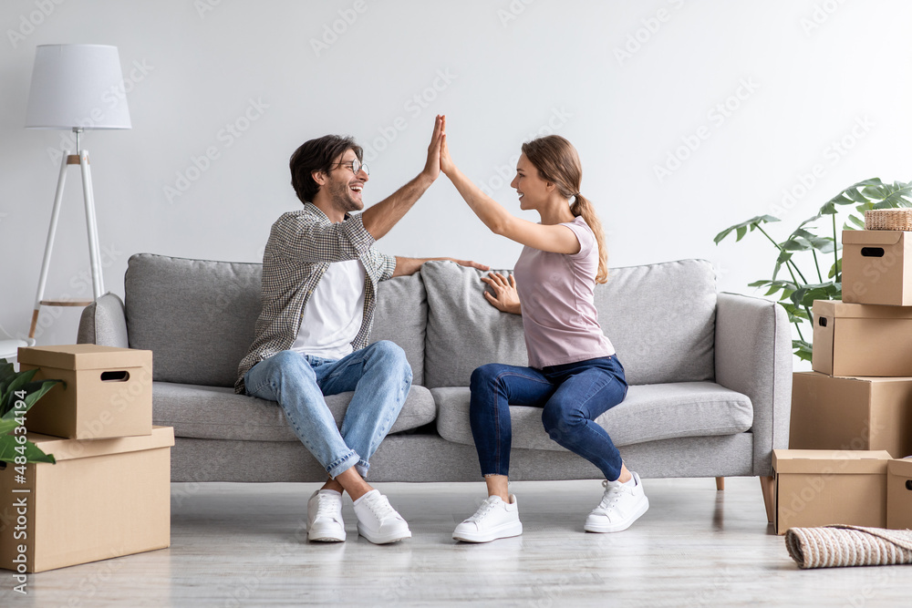 Satisfied european young husband and wife give high five on sofa in living room interior with boxes Satisfied european young husband and wife give high five on sofa in living room interior with boxes