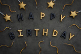 Happy birthday letters in shiny gold and matte black color on a gray background, golden candles in the form of stars and waves