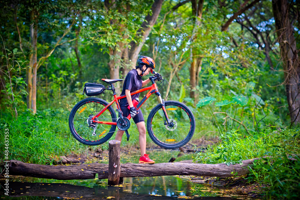woman cyclist in action of carry owns mountain bicycle crossing the small clack of ditch canal in the jungle forest, extreme riding sport trail in the route of competitive area