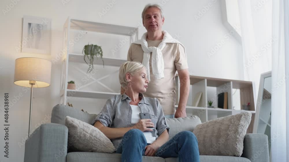 Family secret. Married couple. Trust problem. Casual middle-aged man spying information in mobile phone of wife coming by her sitting sofa in light home room interior. Family secret. Married couple. Trust problem. Casual middle-aged man spying information in mobile phone of wife coming by her sitting sofa in light home room interior.