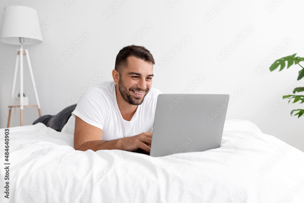 Smiling caucasian attractive millennial male in white t-shirt woke up, chatting, surfing in internet on pc on bed Smiling caucasian attractive millennial male in white t-shirt woke up, chatting, surfing in internet on pc on bed