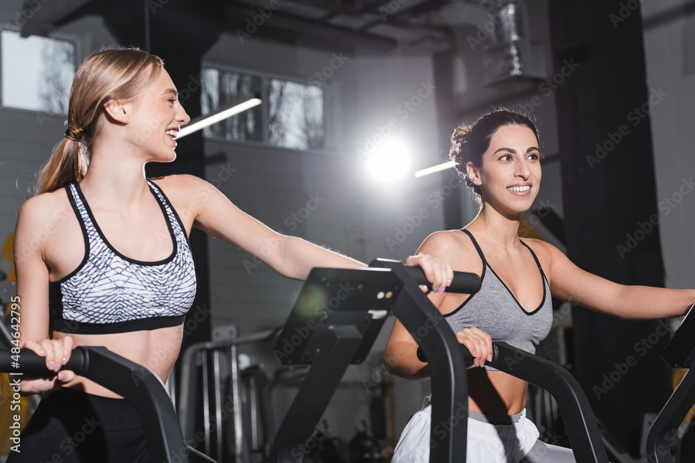 Fototapeta premium Cheerful interracial women training on elliptical trainers in sports center.