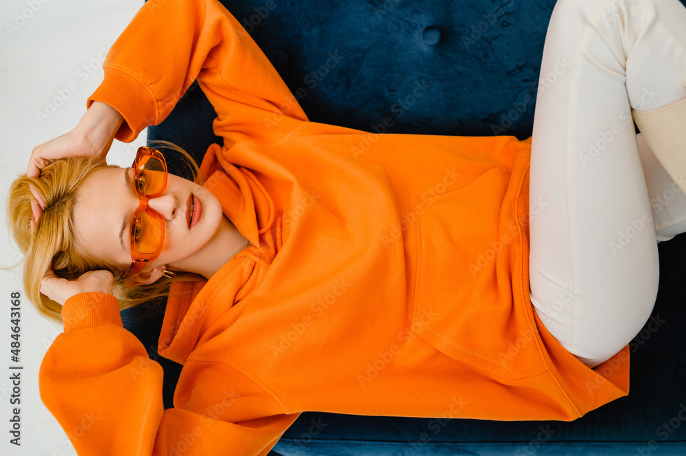 Fashionable confident blonde woman wearing trendy orange sweatshirt, color glasses, posing on blue sofa - obrazy, fototapety, plakaty 