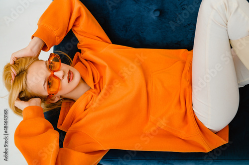 Fashionable confident blonde woman wearing trendy orange sweatshirt, color glasses, posing on blue sofa