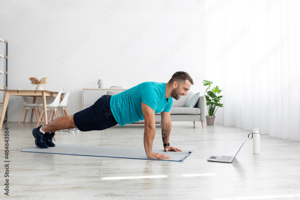 Happy millennial european man doing plank or push ups on mat with laptop in living room interior, profile Happy millennial european man doing plank or push ups on mat with laptop in living room interior, profile