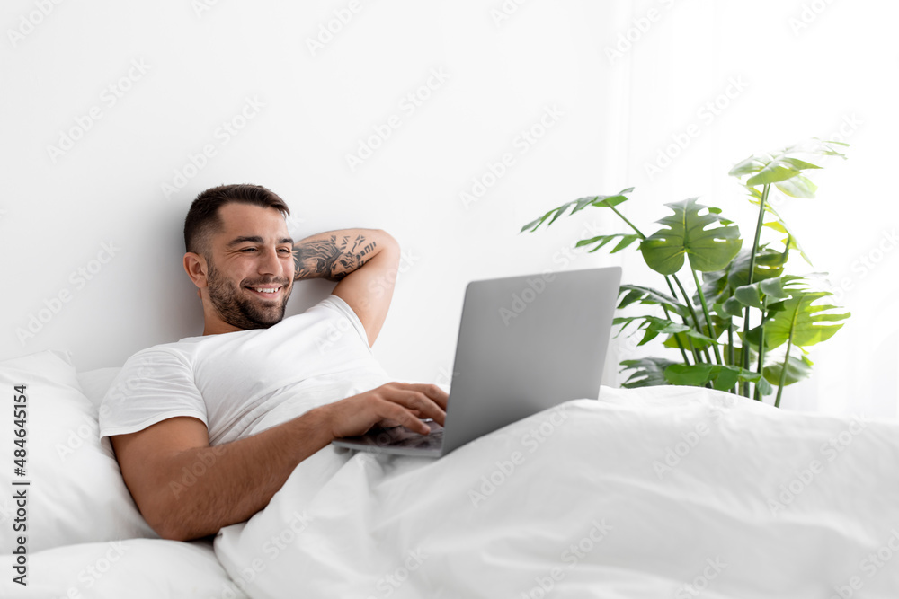 Satisfied young muscular caucasian man typing on computer lies on bed in bedroom interior Satisfied young muscular caucasian man typing on computer lies on bed in bedroom interior
