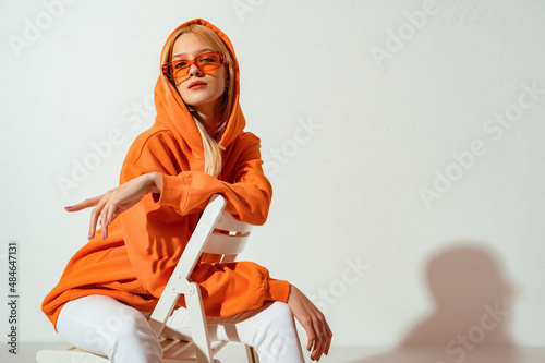 Fashionable confident blonde woman wearing trendy orange sweatshirt, color sunglasses, posing on white background. Copy, empty space for text