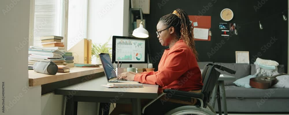Zoom in shot of young African American woman with disability sitting in wheelchair at desk, using laptop and taking notes while working online from home