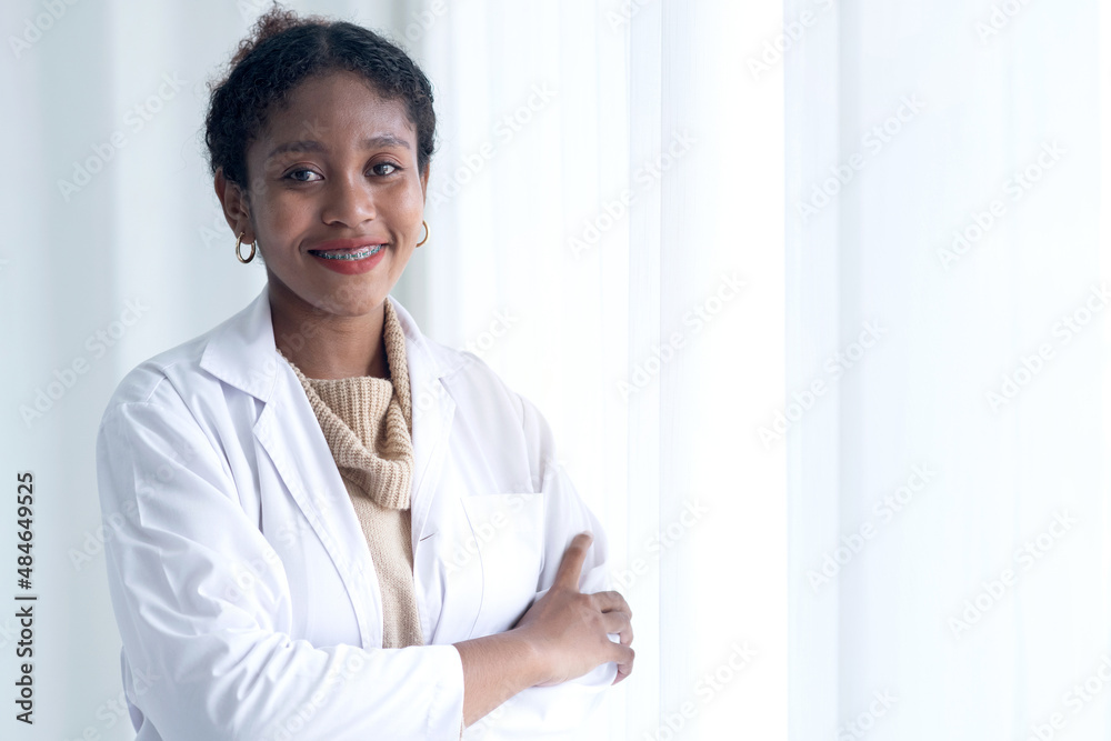 Young African American female doctor with charming smile wears lab coat and holds tablet in hand, stand with arms crossed near the window Young African American female doctor with charming smile wears lab coat and holds tablet in hand, stand with arms crossed near the window