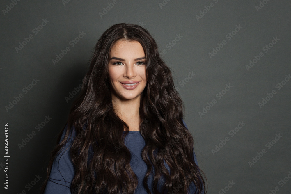Portrait of serious beautiful female with black curly hair, has minimal makeup, looks smiling at camera, stands against gray background Portrait of serious beautiful female with black curly hair, has minimal makeup, looks smiling at camera, stands against gray background