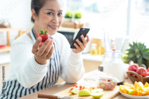 Asian middle age woman show strawberry at her kitchen, and holding a smartphone in hand, prepared fruits and vegetables for breakfast, healthy food at home