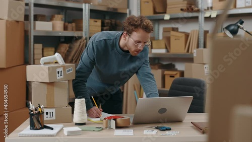 Ginger caucasian man standing at the table, looking at the laptop screen and writing information about parcel delivery while working at the warehouse. Post service and small business concept.