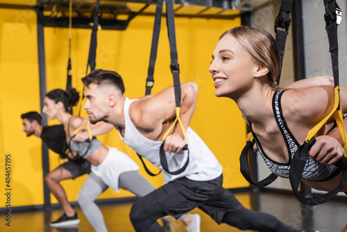 Smiling sportswoman training with suspension straps near blurred friends in gym.