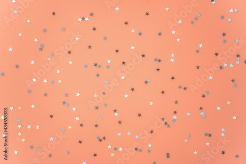 Silver stars glittering confetti on peach color background. Trendy festive holiday backdrop. Many star-shaped particles for a postcard, invitation or web banner