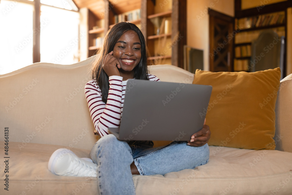Online job, freelancing, remote education. Young black woman sitting on sofa with laptop, working or learning from home