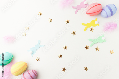 Festive background with paper rabbits and colorful eggs and golden confetti on a white wooden background  top view. Easter card with copy space and decorations.