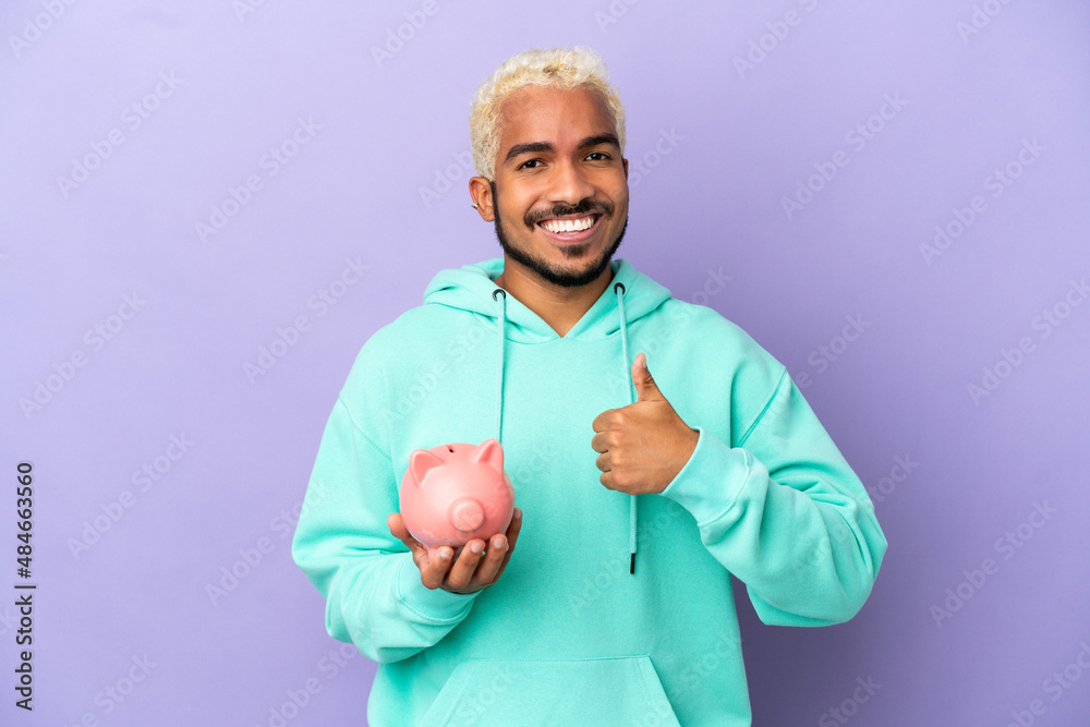 Young Colombian man holding a piggybank isolated on purple background giving a thumbs up gesture Young Colombian man holding a piggybank isolated on purple background giving a thumbs up gesture