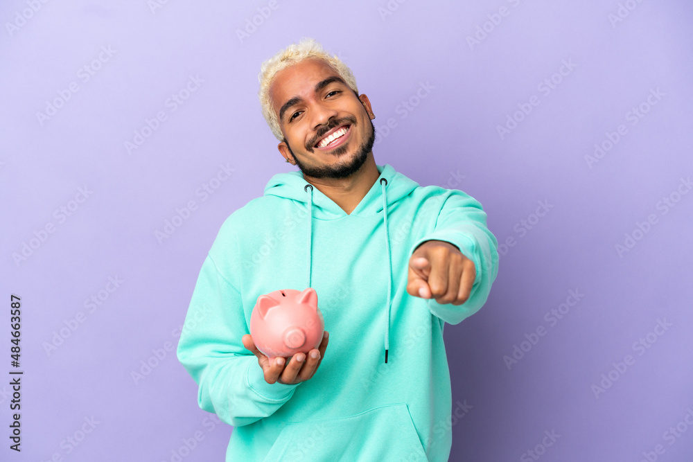 Young Colombian man holding a piggybank isolated on purple background pointing front with happy expression Young Colombian man holding a piggybank isolated on purple background pointing front with happy expression