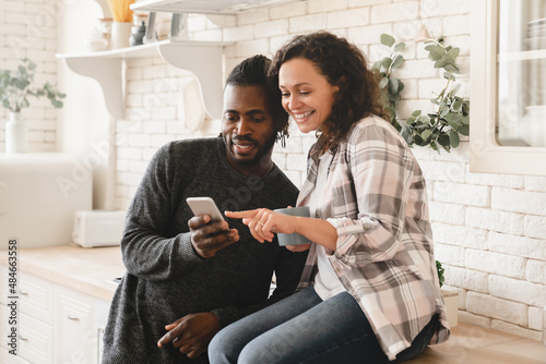 Middle-aged mature happy african couple using smart phone cellphone for surfing social media, sharing photos, checking e-mail, using mobile application online relaxing at home kitchen