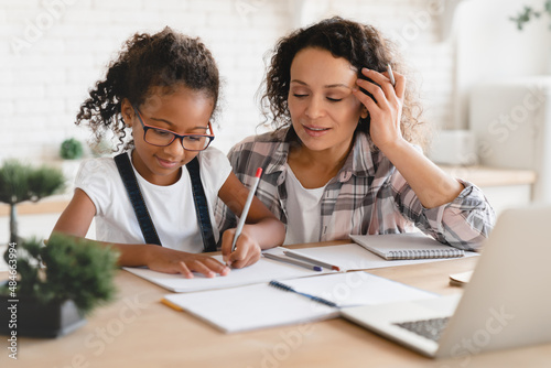 African-american mom mother tutor nanny childminder helping assisting with homework school project to a preteen daughter. Homeschool concept. E-learning on laptop photo
