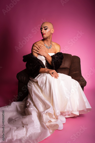 Sad gender fluid transgender gay man wearing wedding dress sitting in armchair on pink background
