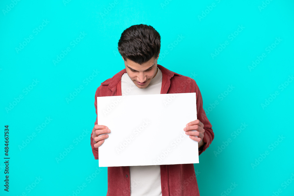 Young handsome caucasian man isolated on blue bakcground holding an empty placard and looking it Young handsome caucasian man isolated on blue bakcground holding an empty placard and looking it