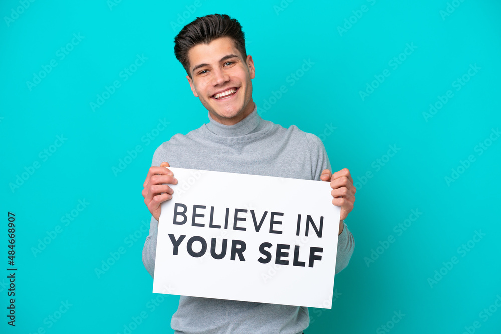 Young handsome caucasian man isolated on blue bakcground holding a placard with text Believe In Your Self with happy expression Young handsome caucasian man isolated on blue bakcground holding a placard with text Believe In Your Self with happy expression