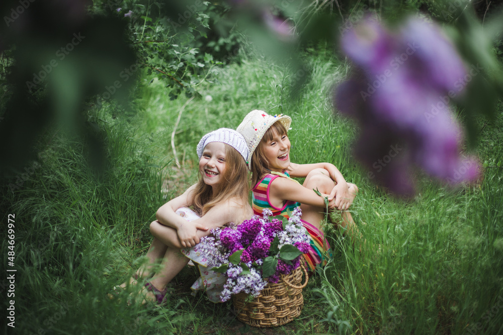 Fototapeta premium Two laughing European girls in white caps sit in a summer garden on the ground with a basket of colorful lilacs.