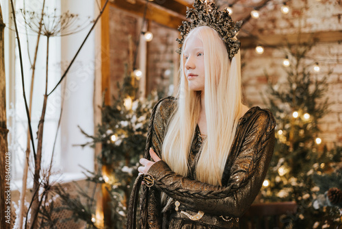 young albino woman with blue eyes and long white hair in beautiful green dress and crown stands in loft room decorated with wooden greenhouse and Christmas trees with twinkle lights, diverse people photo