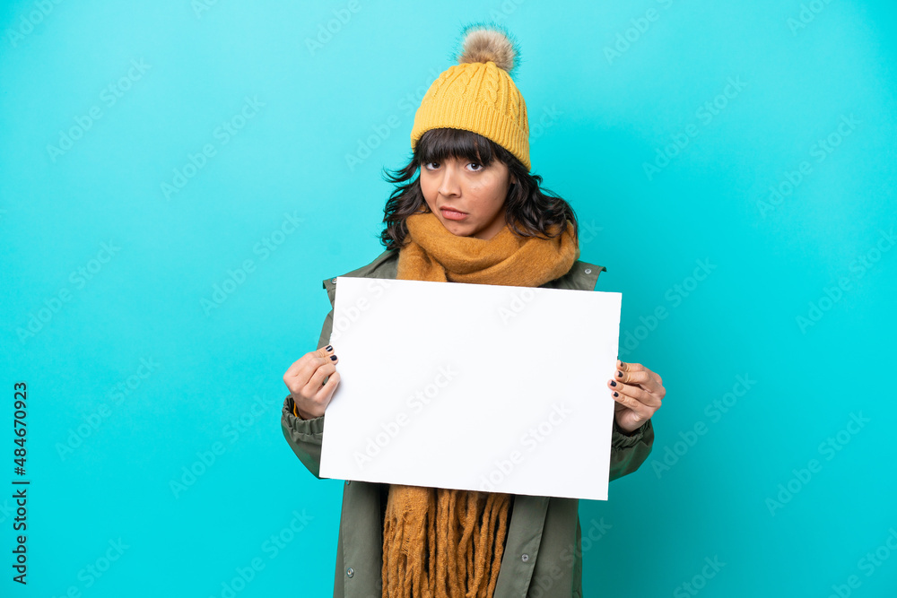 Young latin woman wearing winter jacket isolated on blue background holding an empty placard