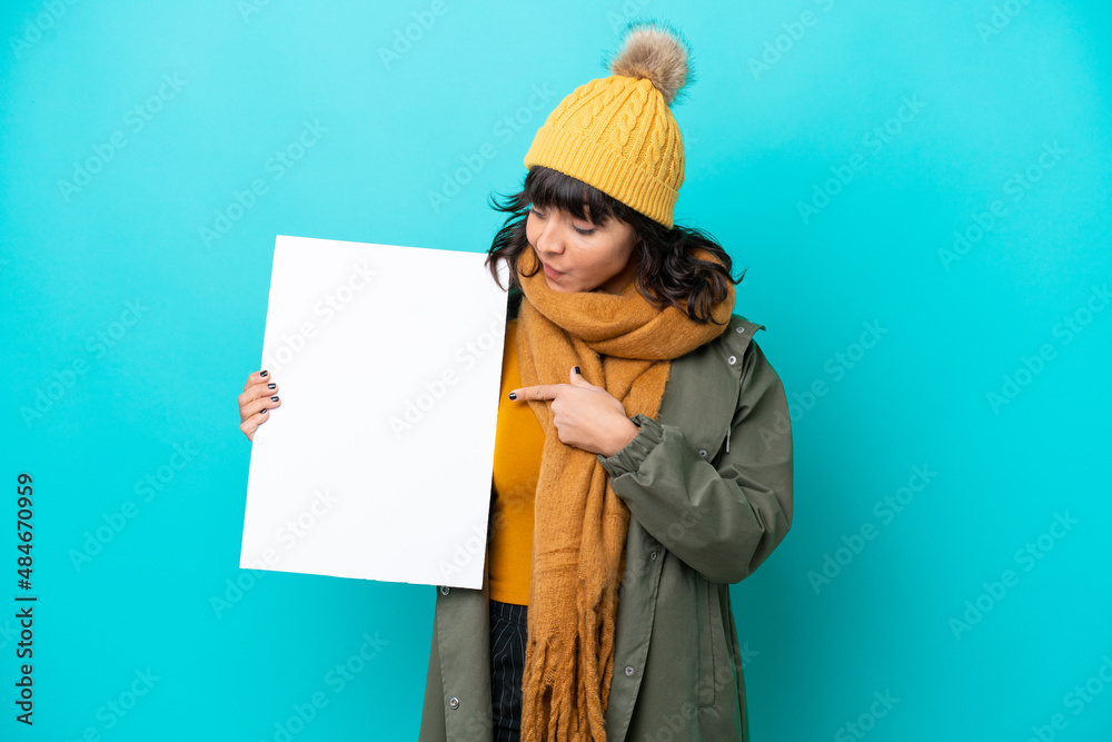 Young latin woman wearing winter jacket isolated on blue background holding an empty placard with happy expression and pointing it Young latin woman wearing winter jacket isolated on blue background holding an empty placard with happy expression and pointing it